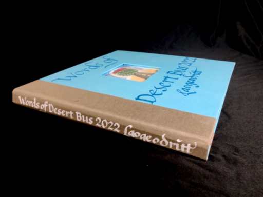 Photograph of a hardbound book. The outer cover is cyan bookcloth, with a desert-brown cloth binding the spine. It is titled 'Words of Desert Bus 2022', author 'Laogeodritt', in hand calligraphy italic script directly on the bookcloth, both on the front cover and spine. In the middle of the front cover, there is a window, revealing a watercolour painting of a round fishhook cactus floating over a straight road in a desert. A white aura around the cactus forms an omega symbol.